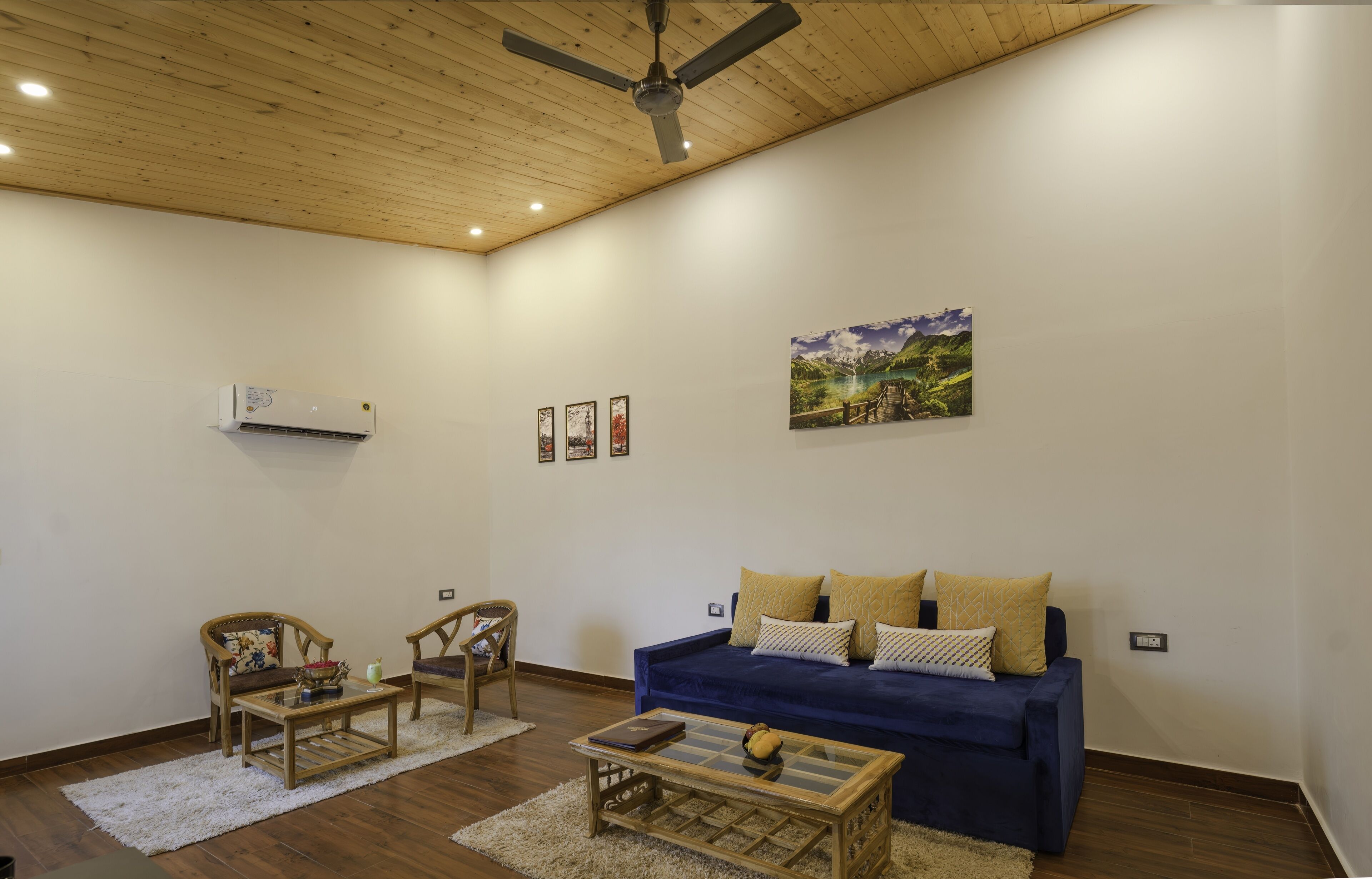 Una sala de estar con un sofá azul, dos sillones, una mesa de centro, un ventilador de techo y un cuadro en la pared.