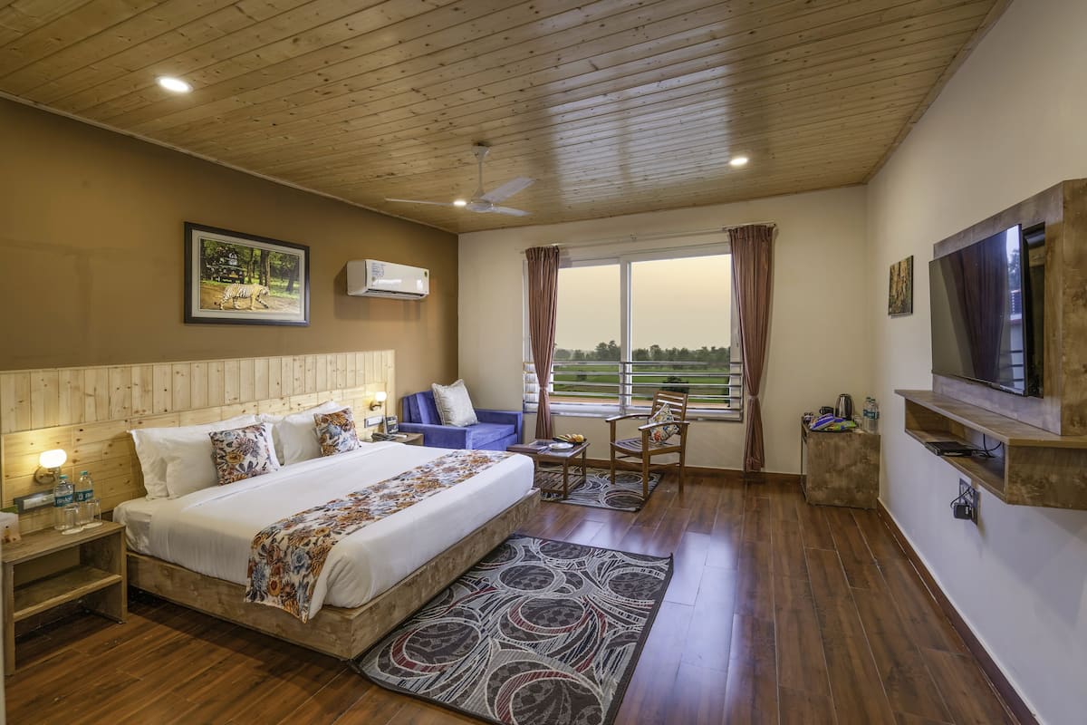 A bedroom with a wooden ceiling, a bed with a floral bedspread, a blue sofa, a TV mounted on the wall, and a window with curtains.