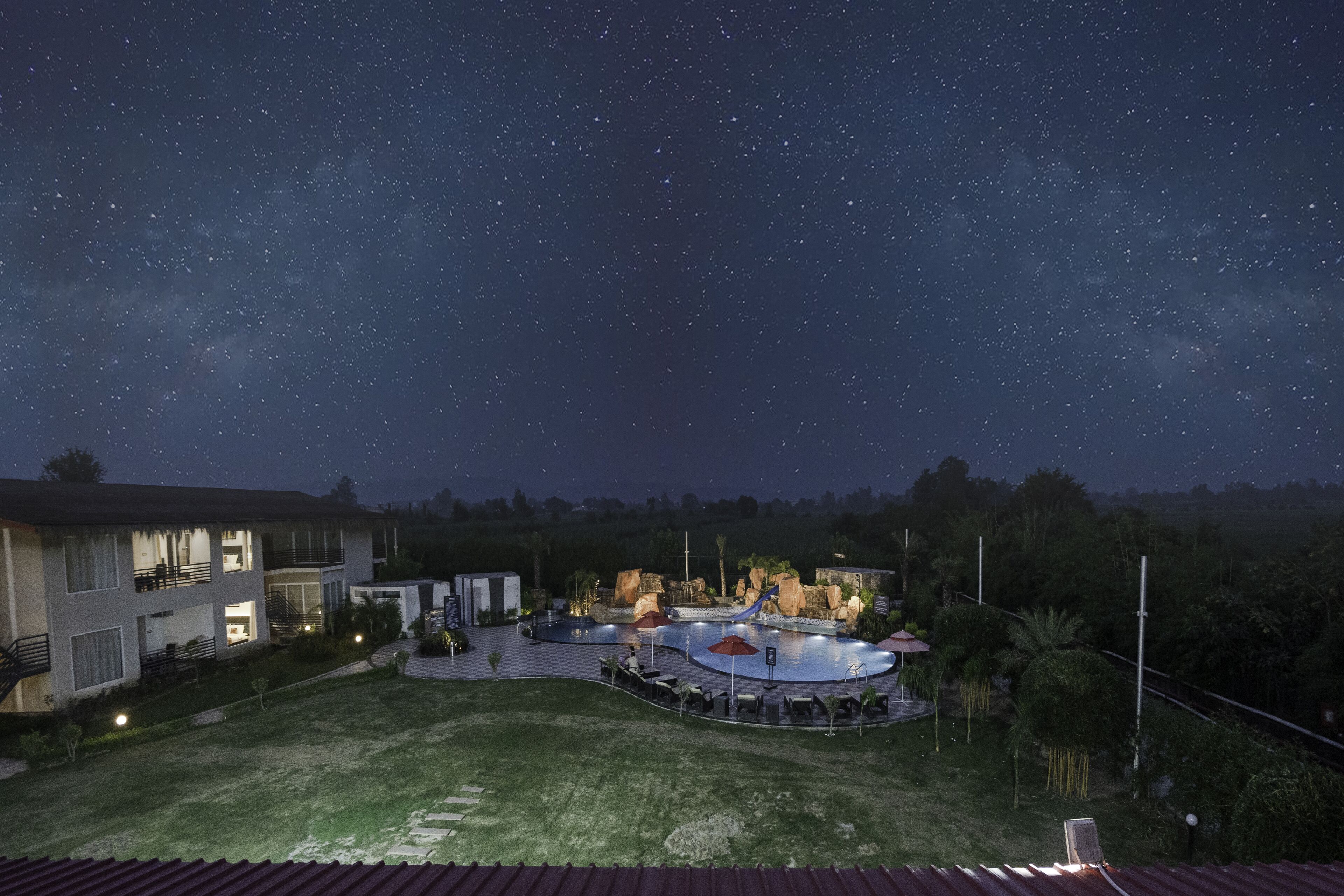 A nighttime view of a residential area with a swimming pool, outdoor seating, and a starry sky.