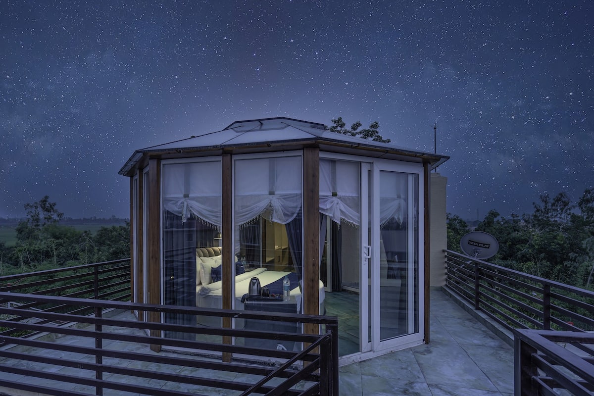 A starry night sky with a glass-roofed gazebo-style room featuring a bed and a small table.