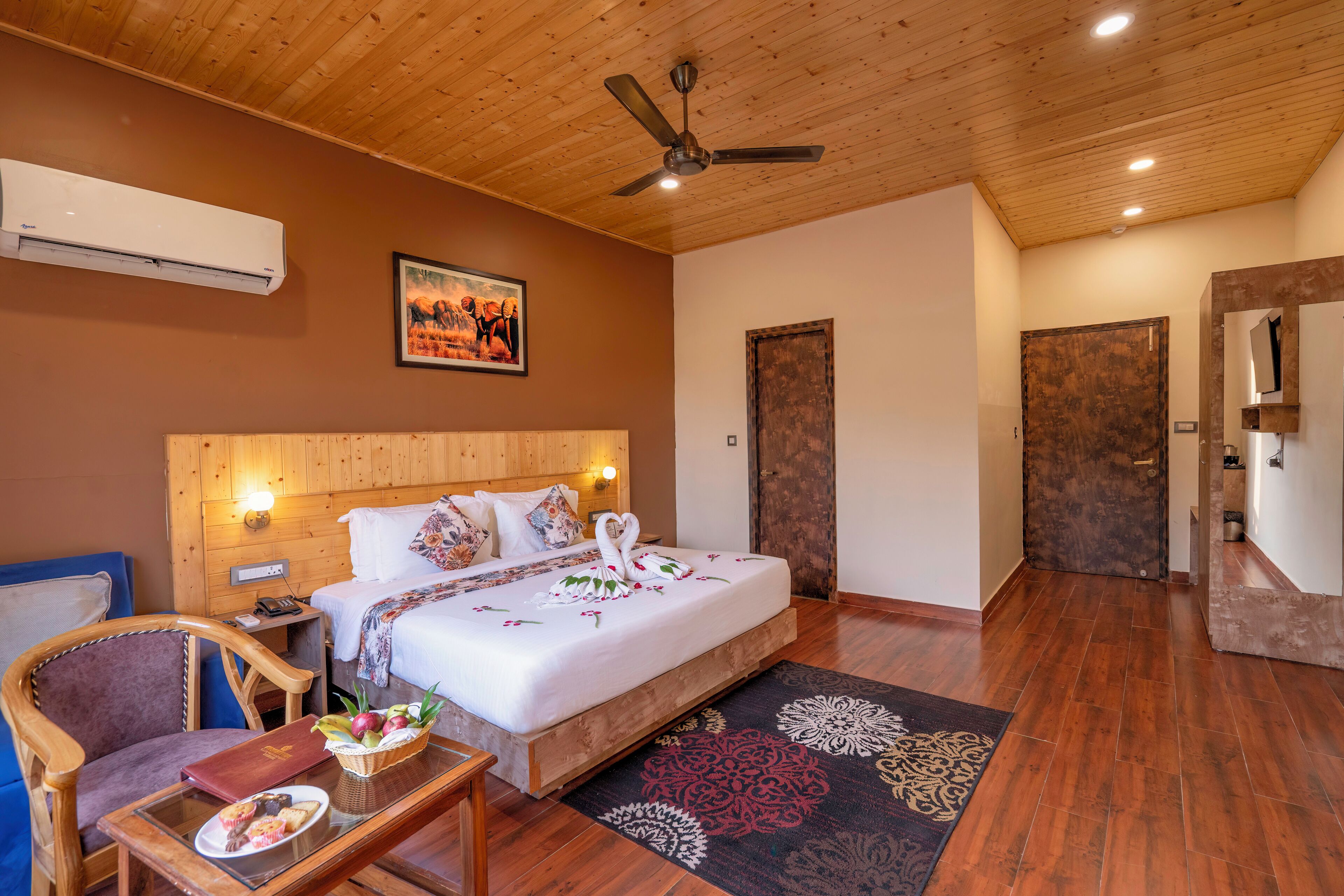 A hotel room with a wooden ceiling, a bed with a white duvet and decorative swan pillows, a wooden nightstand, a chair, a small table with a basket of fruit, and a patterned rug.