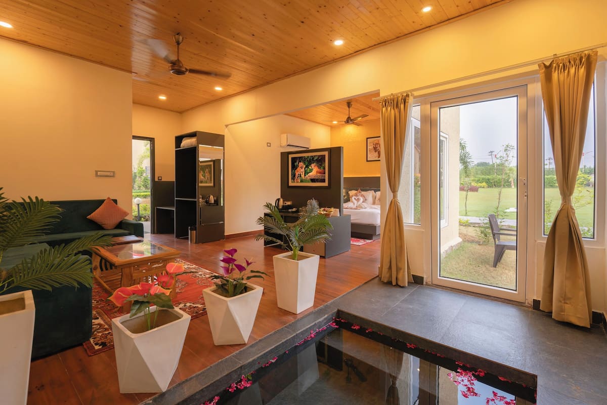 A modern living room with a sofa, coffee table, and potted plants. There is a sliding glass door leading to a balcony with a view of the garden.
