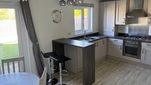 A modern kitchen with a central island, stainless steel appliances, and a dining area with a round table and bar stools. - Remarkable 3-bed House in Wirral (Wirral)