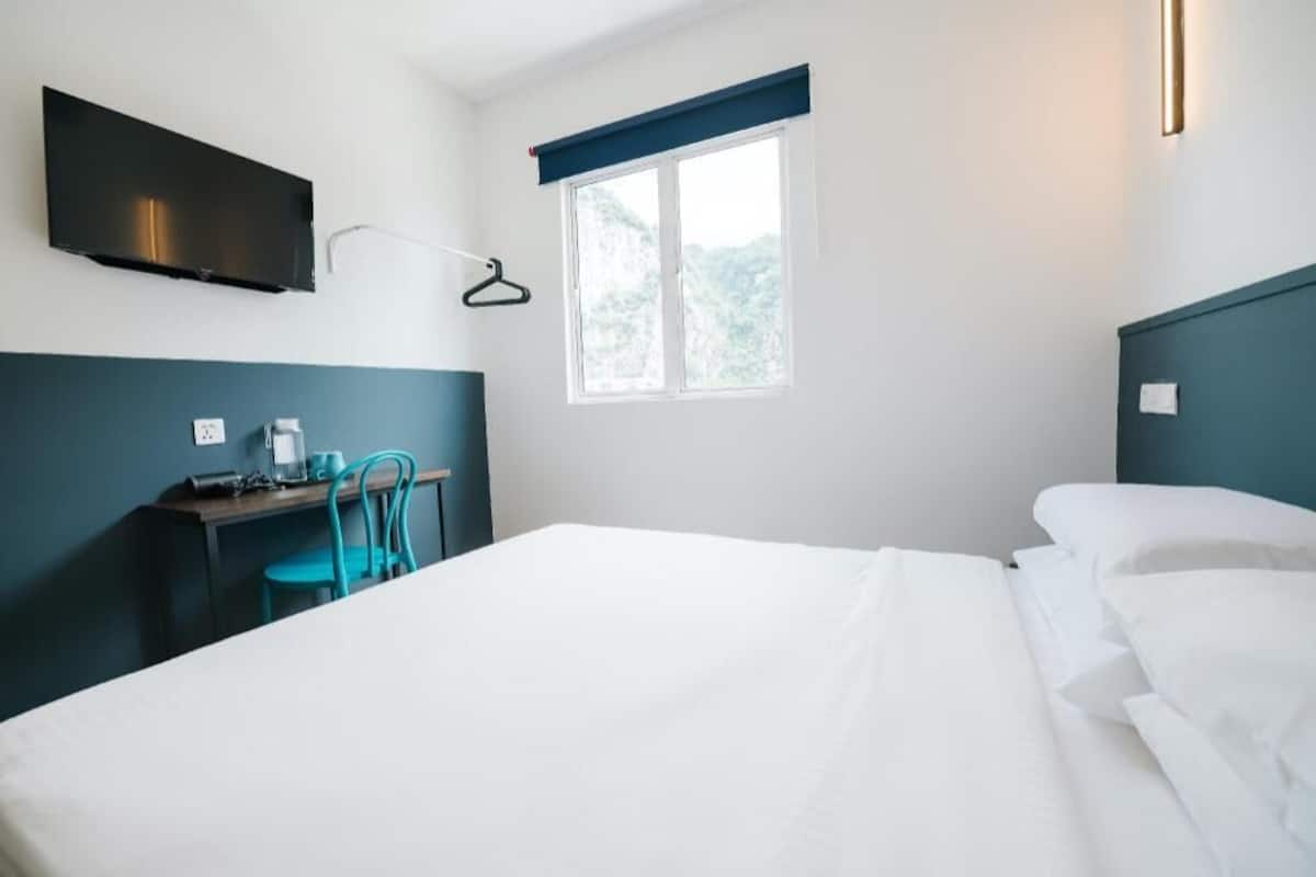Hotellihuone, jossa on sänky, työpöytä tuolilla, seinälle asennettu televisio ja ikkuna, josta avautuu näkymä vehreään maisemaan.
