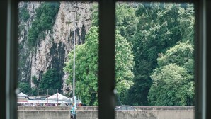 A view through a window of a concrete barrier with a yellow car on the road, a blue tent-like structure, and a rocky cliff with dense green foliage in the background. - The Concept Hotel KL - Batu Caves (Batu Caves)