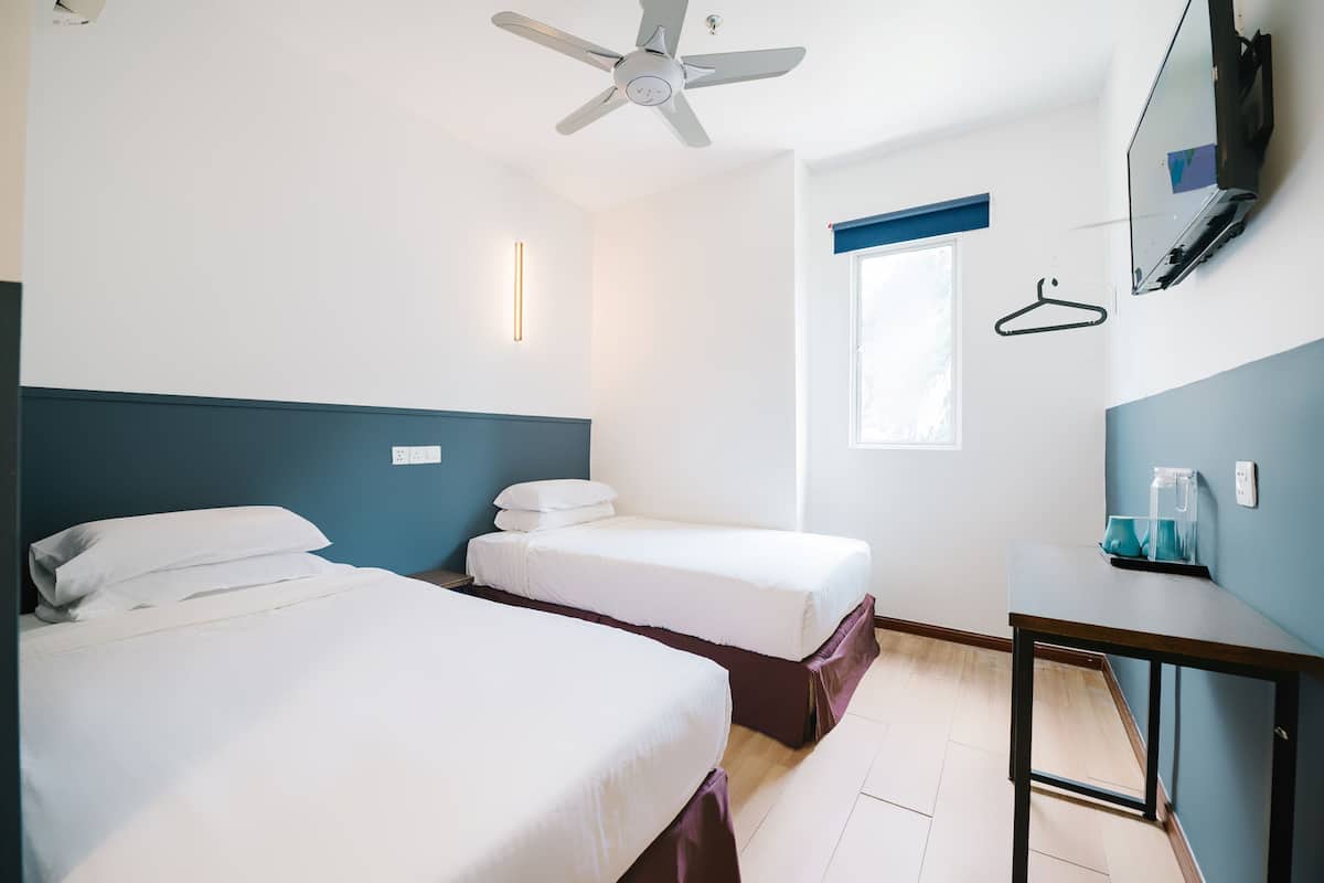 Hotellihuone, jossa on kaksi sänkyä, kattotuuletin, työpöytä, peili ja sinisellä kehyksellä varustettu ikkuna.