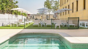 Una piscina con borde de azulejos, rodeada por una valla, y mobiliario de exterior con sombrillas.