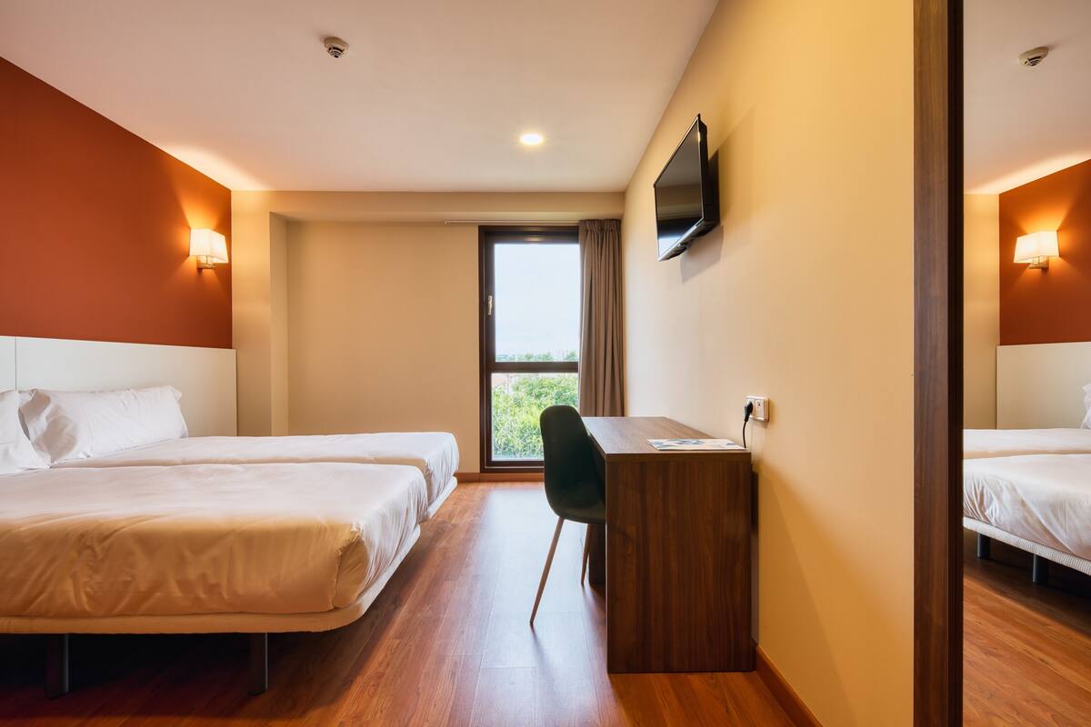 A hotel room with two beds, a desk, a chair, a TV mounted on the wall, and a window with a view of greenery.