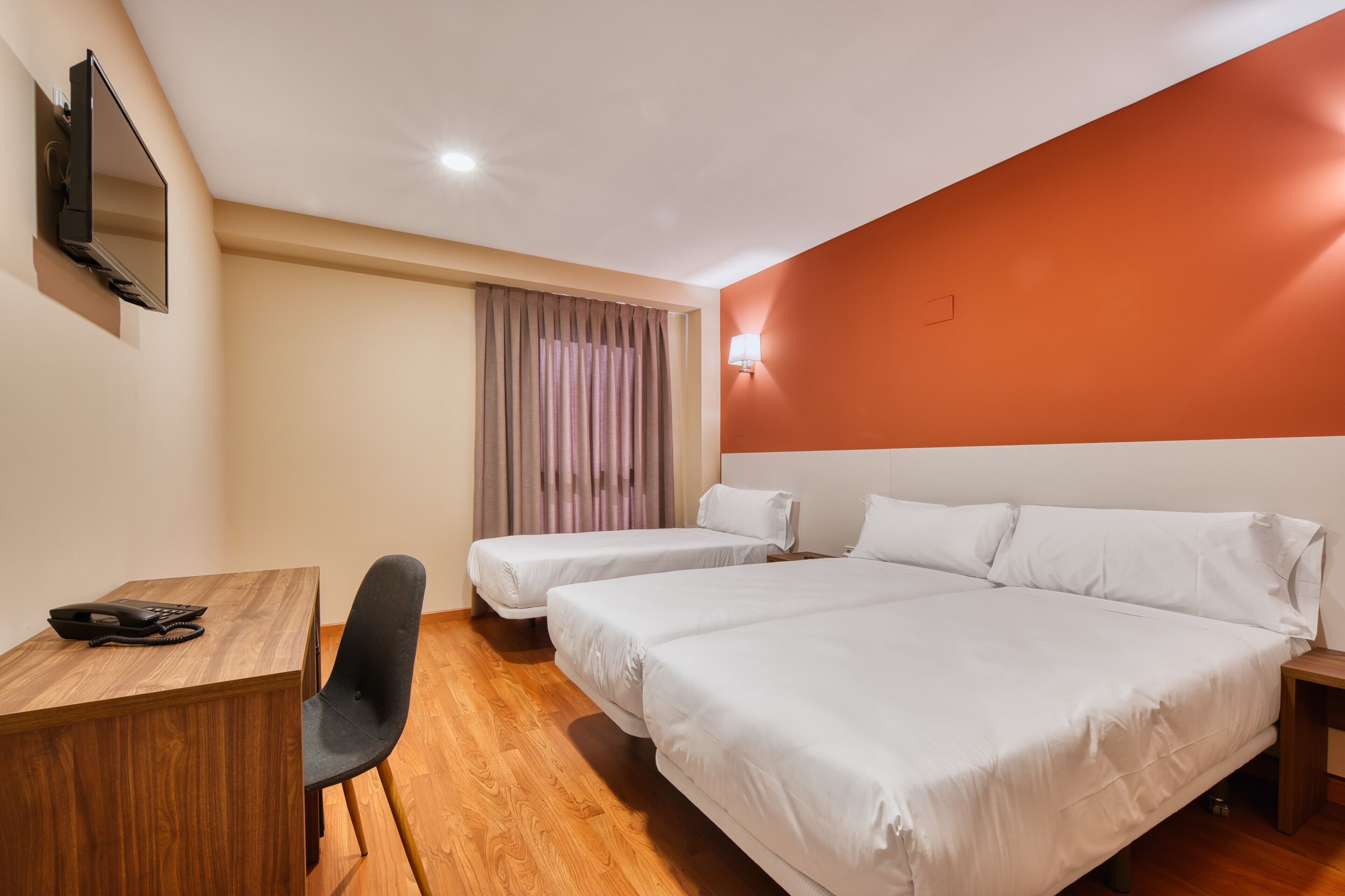 A hotel room with two beds, a desk, a chair, a TV, and a window with curtains.