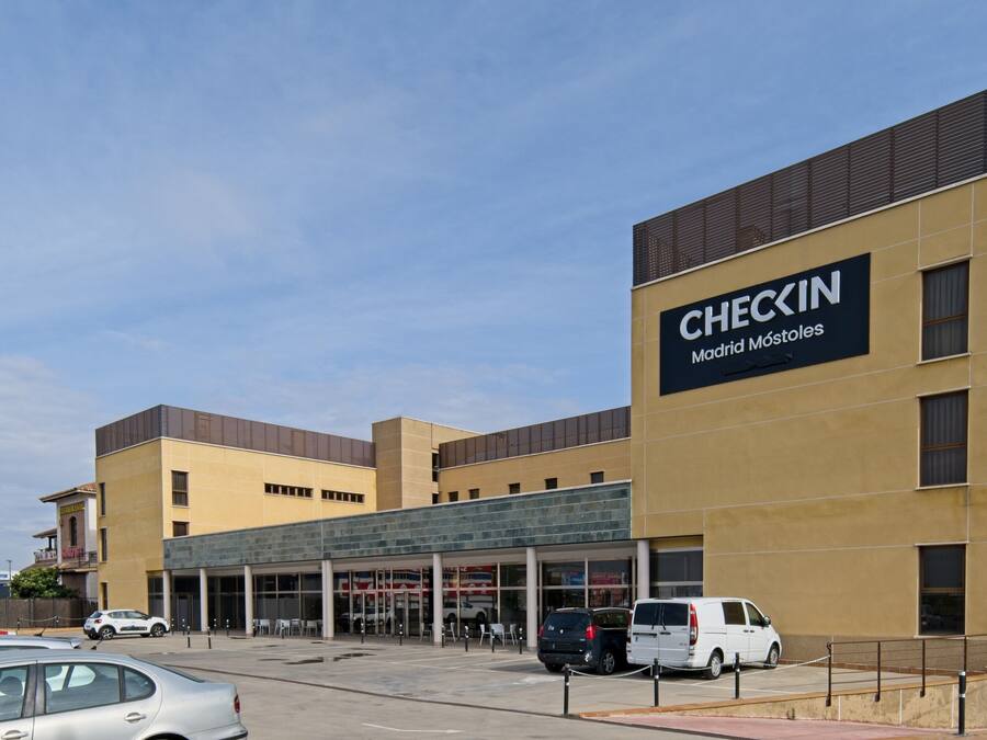 A modern building with a large CHECKIN sign, featuring a glass entrance and a parking area with several cars.
