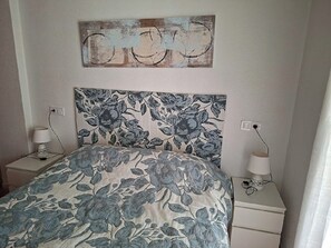 Ein Schlafzimmer mit Blumenmuster-Bettdecke, schlichtem Kopfende, Nachttischlampen und einem an der Wand hängenden Kunstwerk.