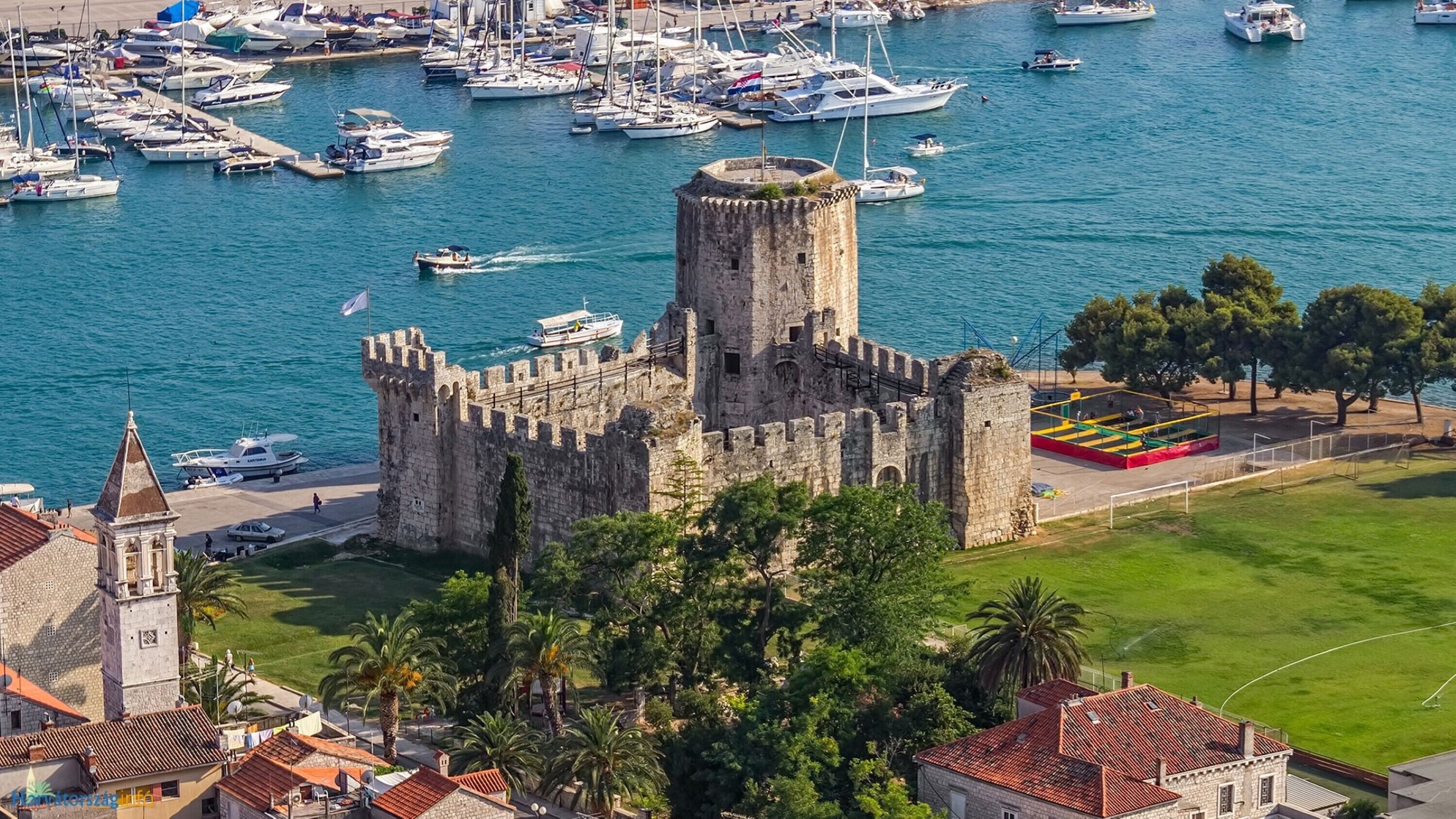 A historic castle with a tower, surrounded by a marina with boats, palm trees, and a sports field.