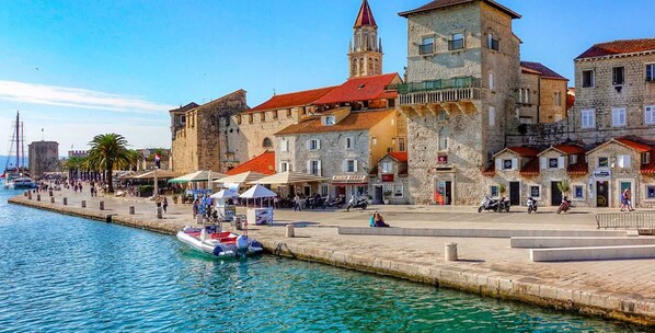 A waterfront promenade with stone buildings, a tower, and a small boat docked at the pier.