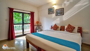 A hotel room with a bed, a nightstand, a window with red curtains, and a view of greenery outside.