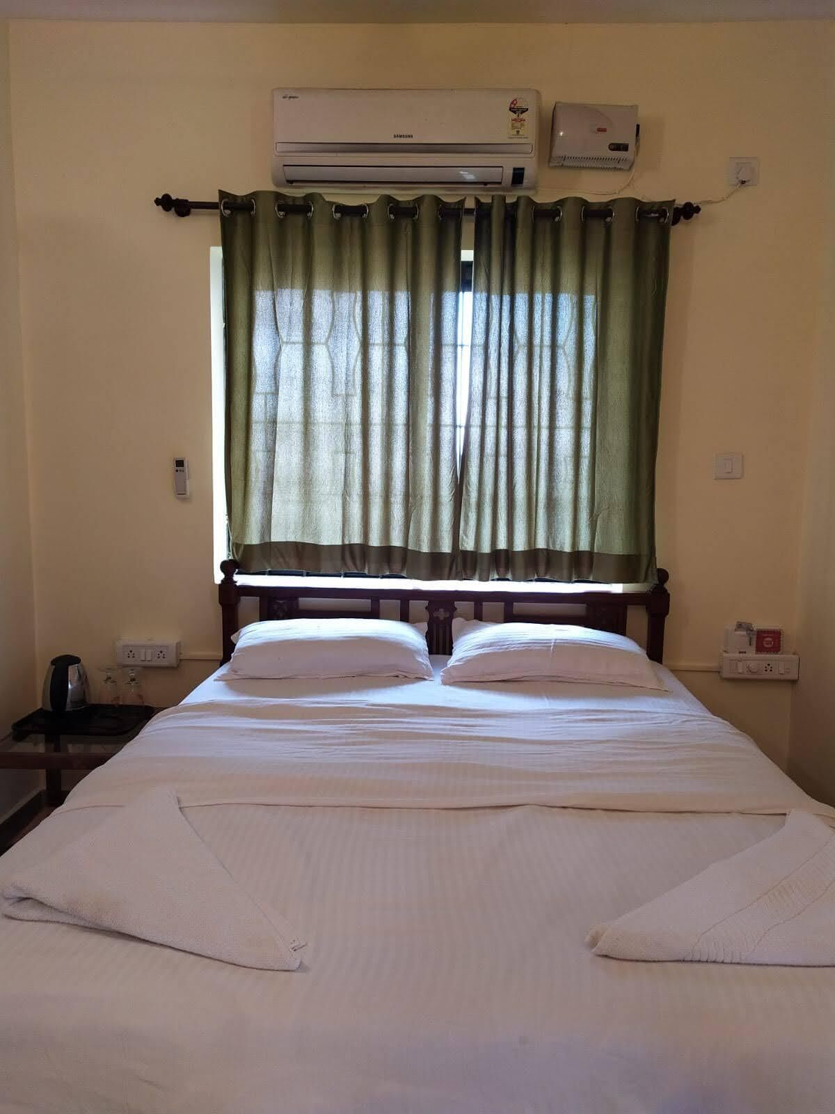 A hotel room with a bed, a nightstand, an air conditioning unit, and a window with green curtains.