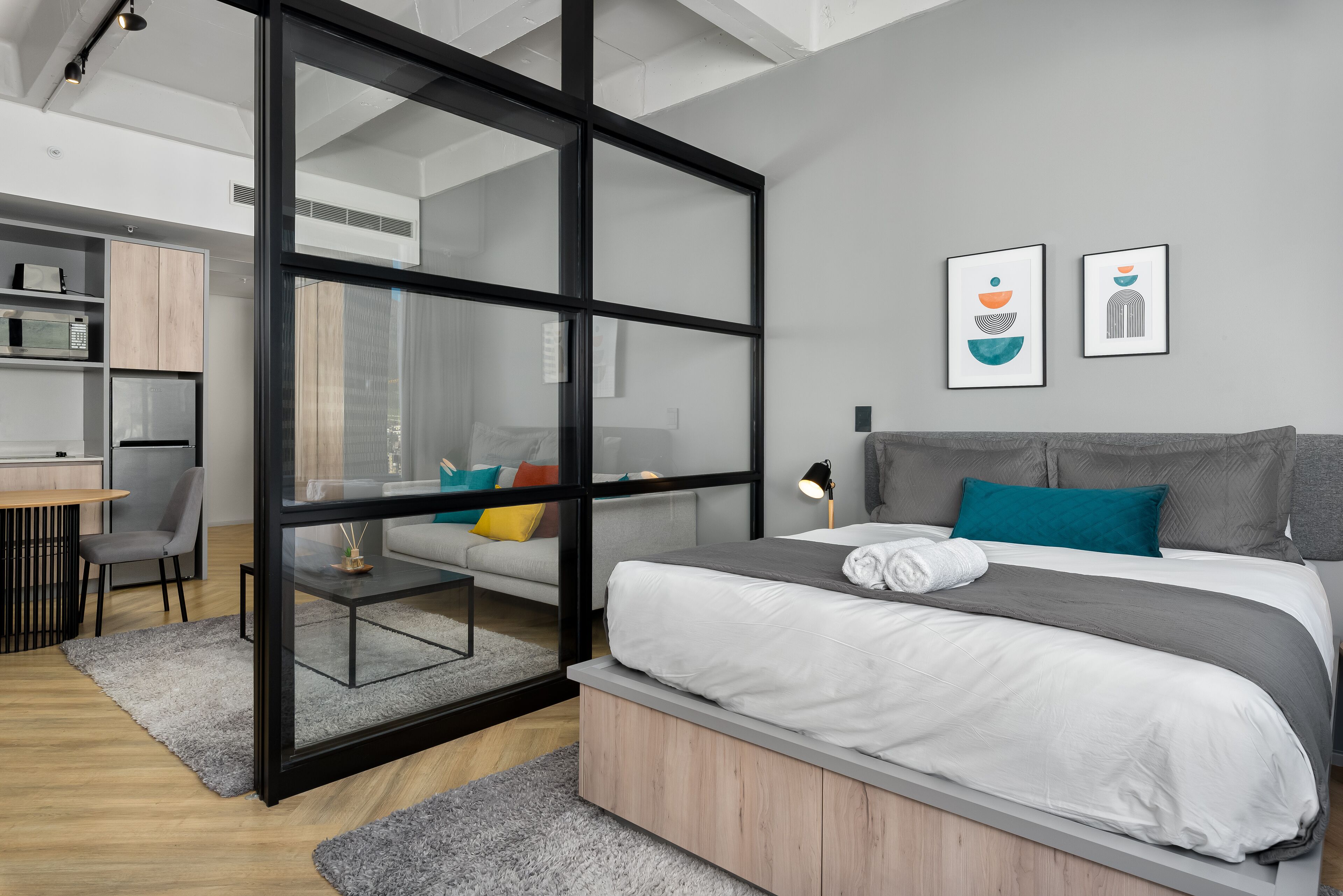 Quarto moderno com divisória de vidro, cama com roupa de cama cinza e branca, escrivaninha com cadeira e área de cozinha compacta.