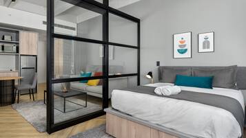 Quarto moderno com divisória de vidro, cama com roupa de cama cinza e branca, escrivaninha com cadeira e área de cozinha compacta.