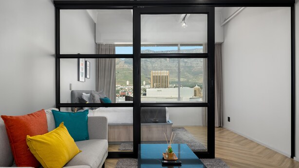 A modern living room with a grey sofa, colorful cushions, a black coffee table, and a large window offering a view of buildings and mountains. - One Thibault 1628 (Cape Town)