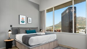 A modern bedroom with a large bed, a nightstand, a lamp, and a view of a cityscape through a large window. - One Thibault 1628 (Cape Town)