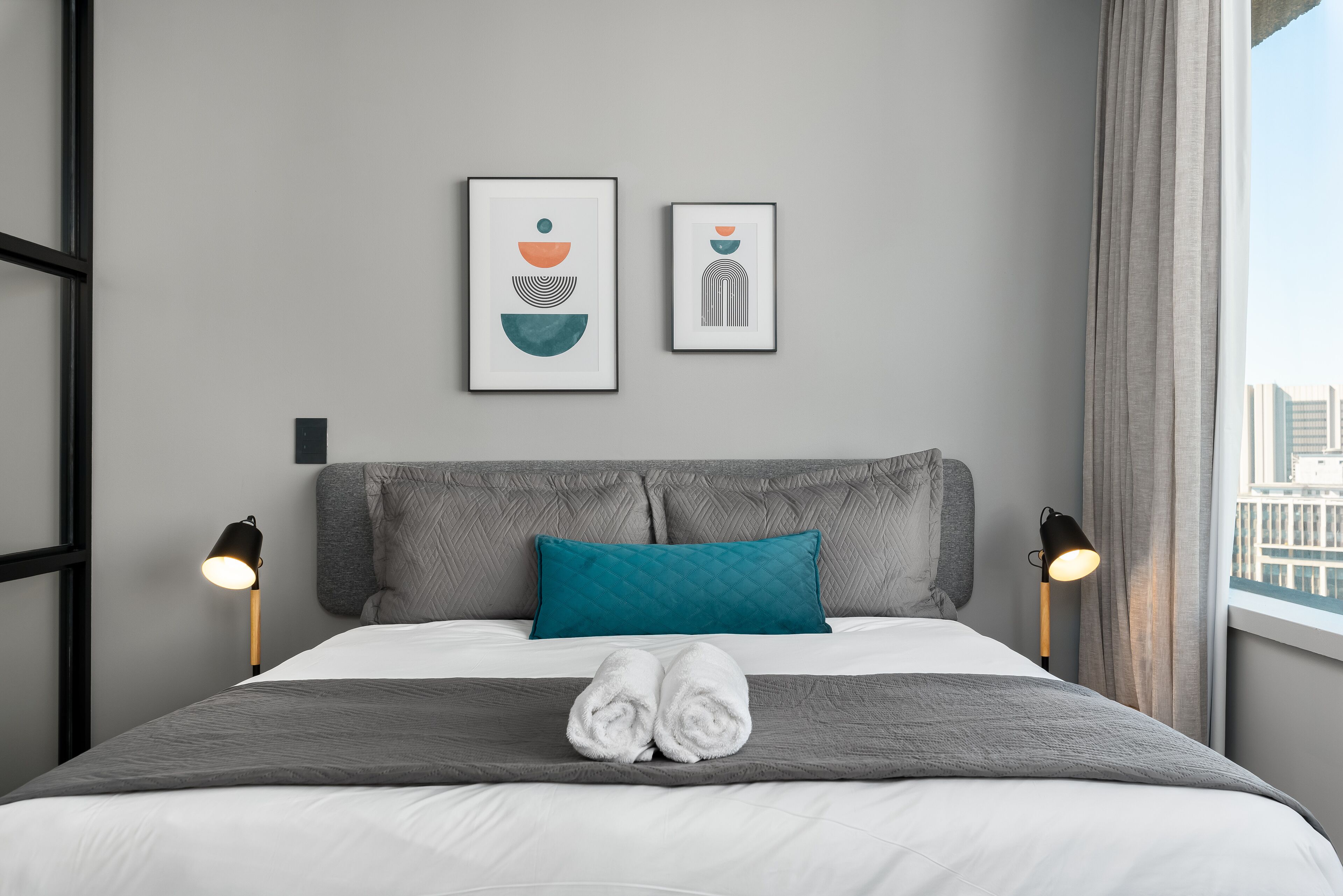A neatly made bed with a grey headboard, two framed artworks above it, and a window with curtains on the right.