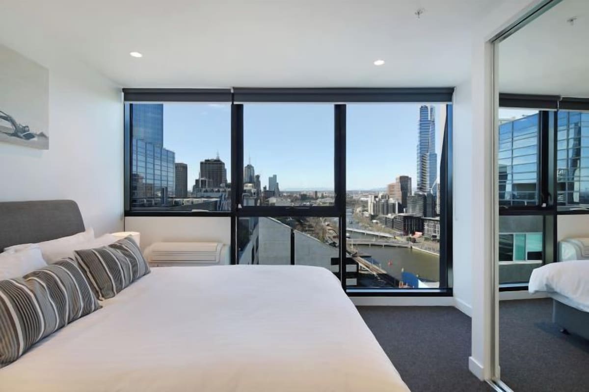 Une chambre d’hôtel dotée d’un grand lit, offrant une vue sur le paysage urbain grâce à de larges fenêtres du sol au plafond, et présentant un design moderne.