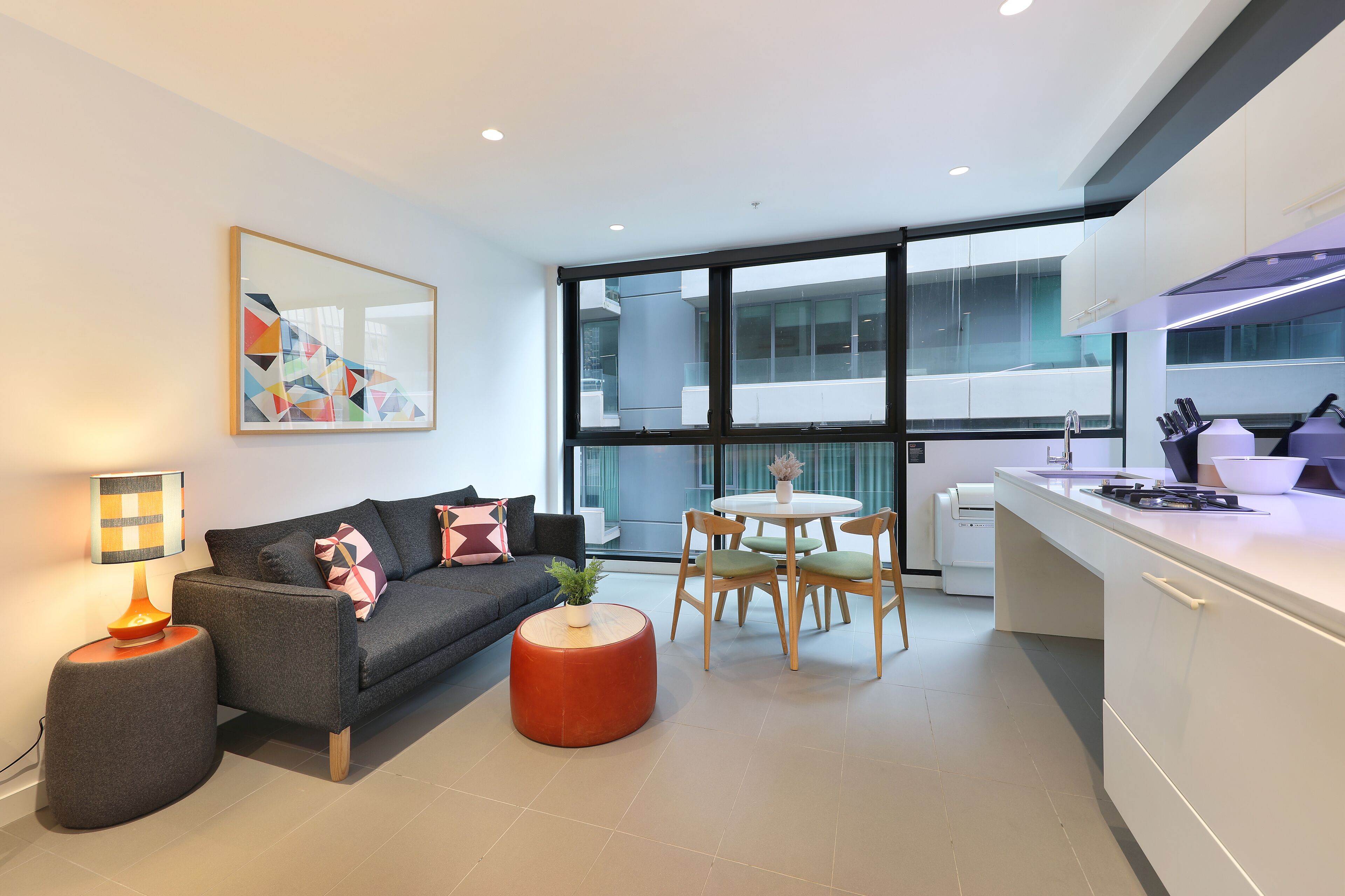 A modern kitchen with a dining area, large windows, and a sofa with a lamp.