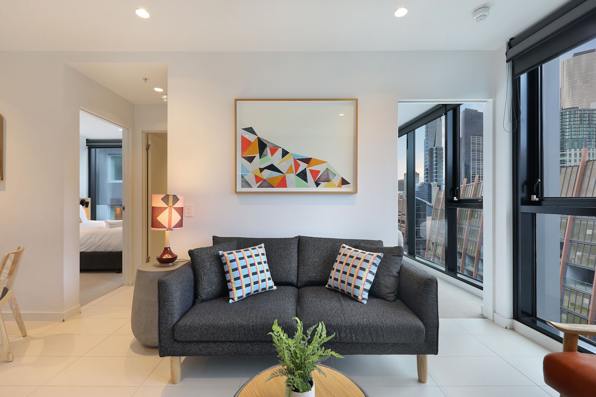 A modern living room with a grey sofa, two patterned throw pillows, a geometric abstract painting, and a view of city buildings through large windows.