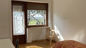 A room with a wooden floor, a bed with a floral bedspread, a desk with a chair, a window with a view of greenery, and a hanging light fixture. - Spacious rooms in the center of Rome (Roma)