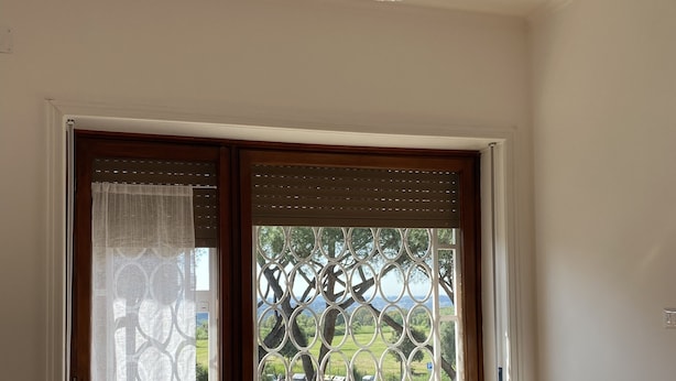 A room with a wooden floor, a wooden chair, a window with a view of trees and a parking lot, and a hanging light fixture. - Spacious rooms in the center of Rome (Roma)