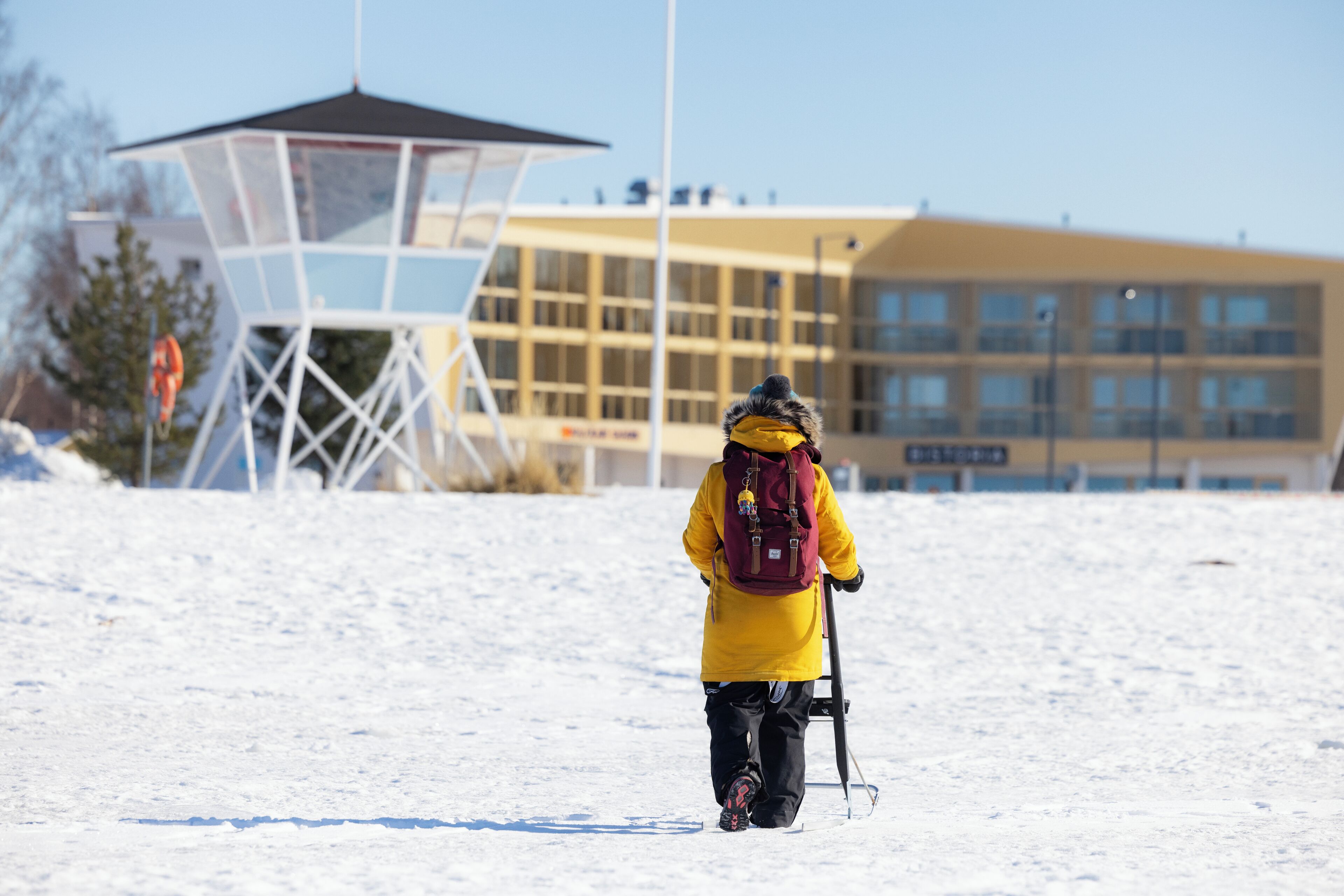 Una persona con una chaqueta amarilla caminando en una zona nevada, con un edificio al fondo.