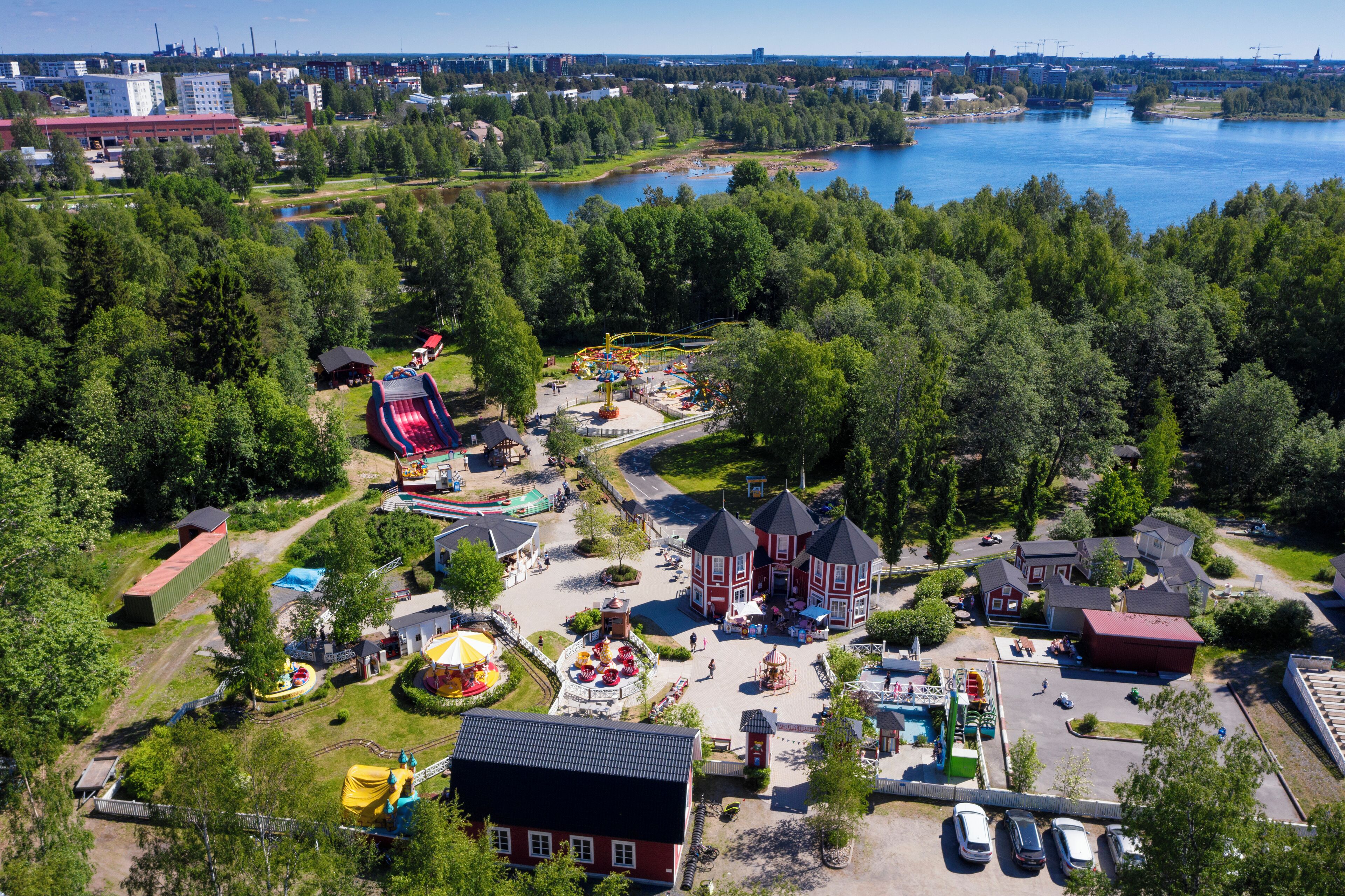 Vista aérea de un parque con diversas atracciones, incluyendo un carrusel, áreas de juego y una gran masa de agua al fondo.