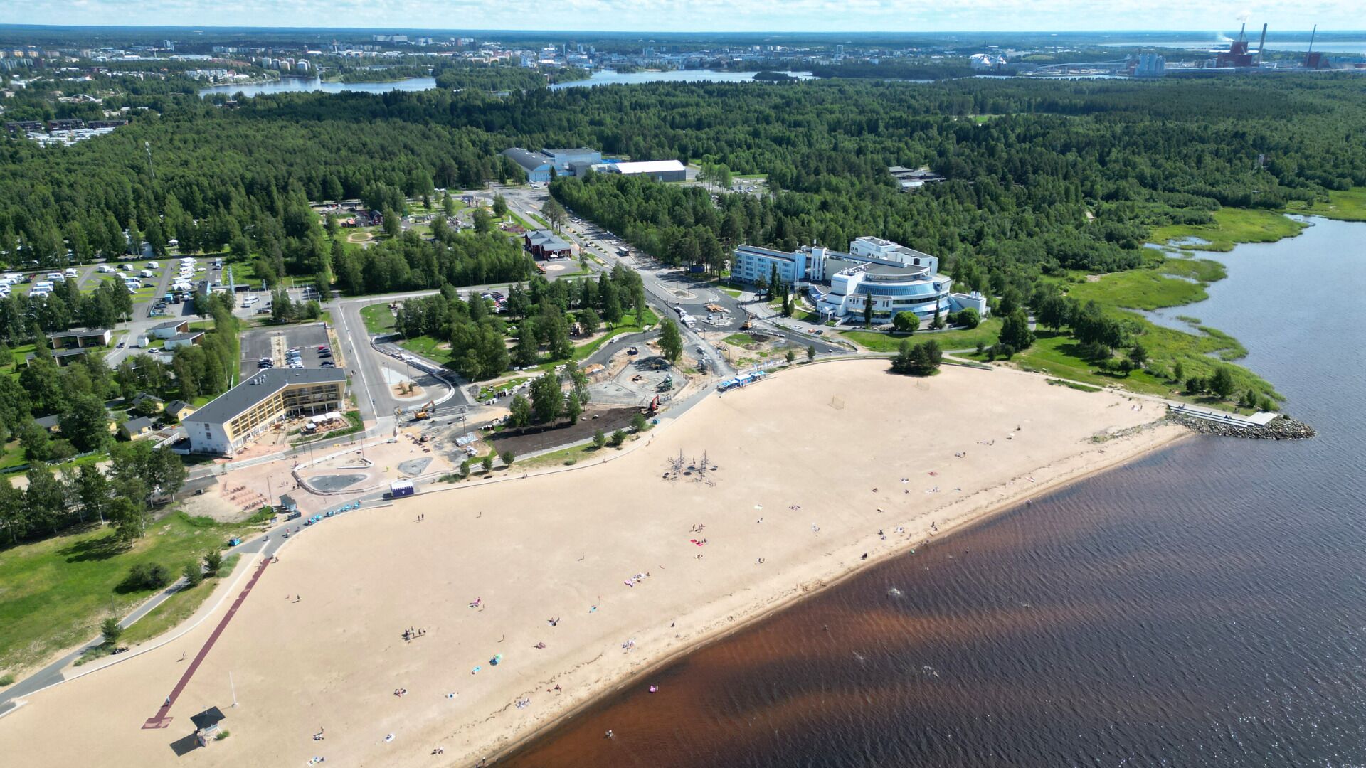 Vista aérea de una zona costera con playa arenosa, estacionamiento y una combinación de edificios residenciales y comerciales.