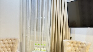 A room with two beige armchairs, a round glass table with four water bottles, and a flat-screen TV on the wall. - ALEXANDER HOTEL (Samarkand)