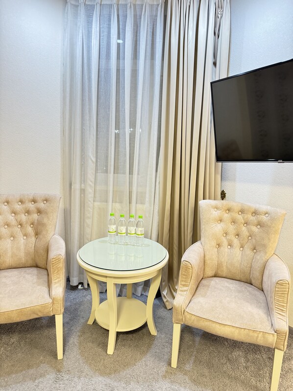 Une pièce avec deux fauteuils beiges, une table ronde en verre sur laquelle sont posées quatre bouteilles d’eau, et un téléviseur à écran plat fixé au mur.
