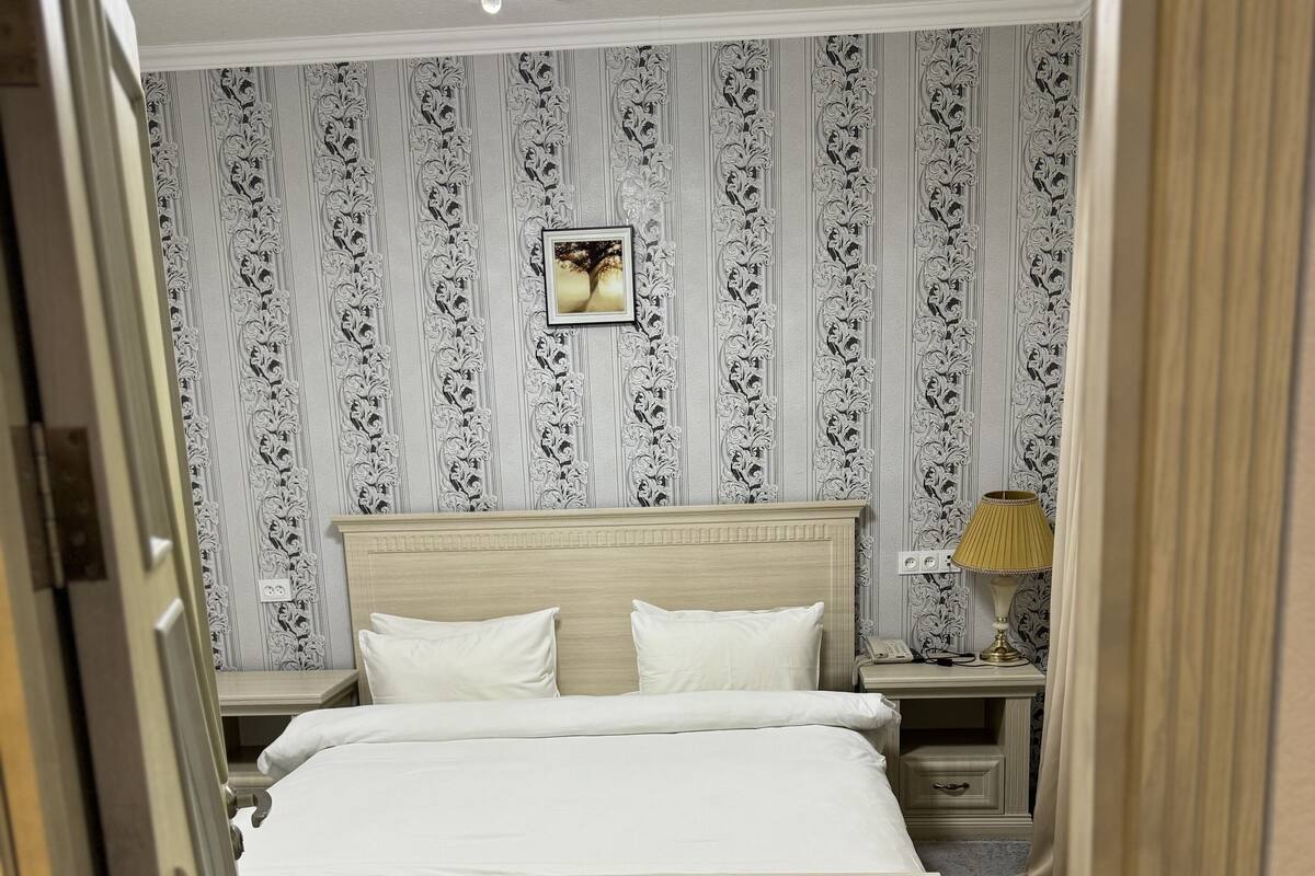 A bedroom with a large bed, a nightstand with a lamp, and a wall with patterned wallpaper.