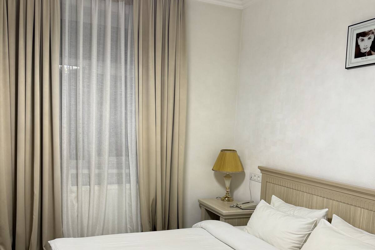 A hotel room with a bed, curtains, a lamp, a bedside table, and a framed picture on the wall.