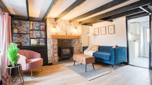 A living room with exposed beams, a brick fireplace, a blue sofa, a pink armchair, and a small table with a plant. - The Cottage (Poulton-le-Fylde)