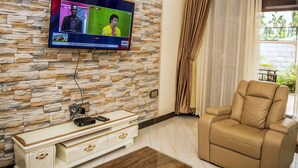 Una sala de estar con pared de piedra, un televisor de pantalla plana montado sobre un mueble de madera, un sofá de cuero color beige y una ventana con cortinas.