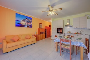 A living room with a sofa, dining table, chairs, a ceiling fan, and a kitchen area with cabinets and appliances.