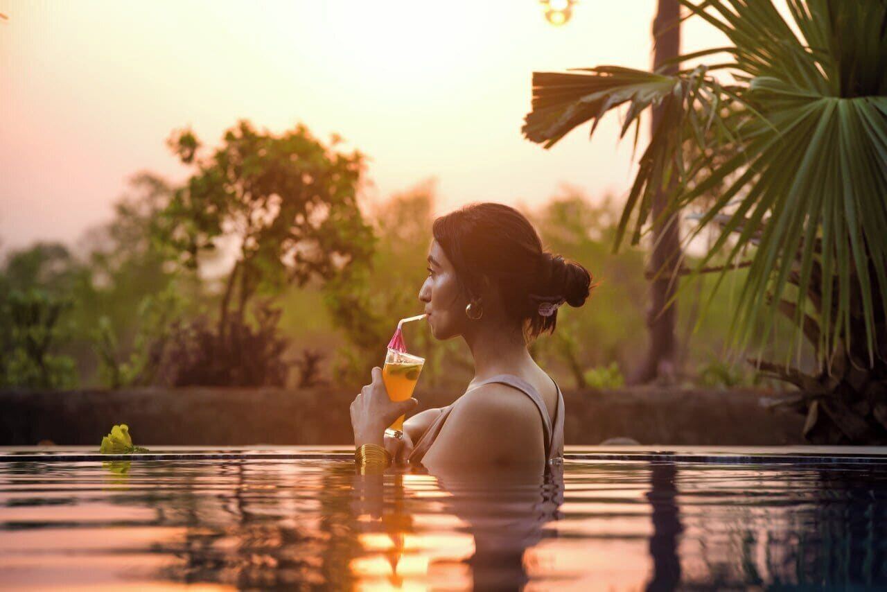 A person in a pool with a drink, surrounded by greenery and a sunset.