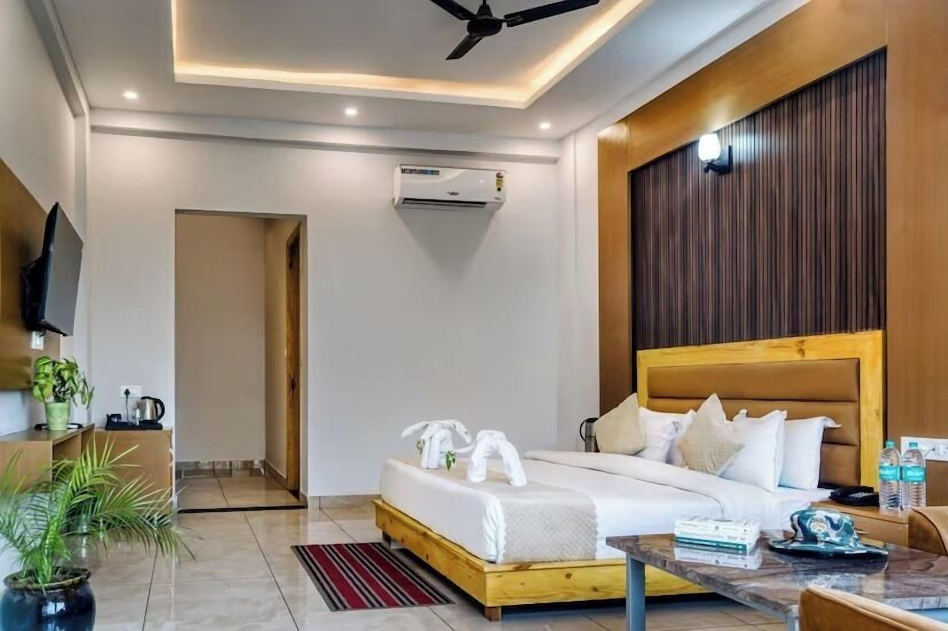 A modern hotel room with a wooden headboard, a flat-screen TV, a bed with white linens, a bedside table with a lamp, and a potted plant.