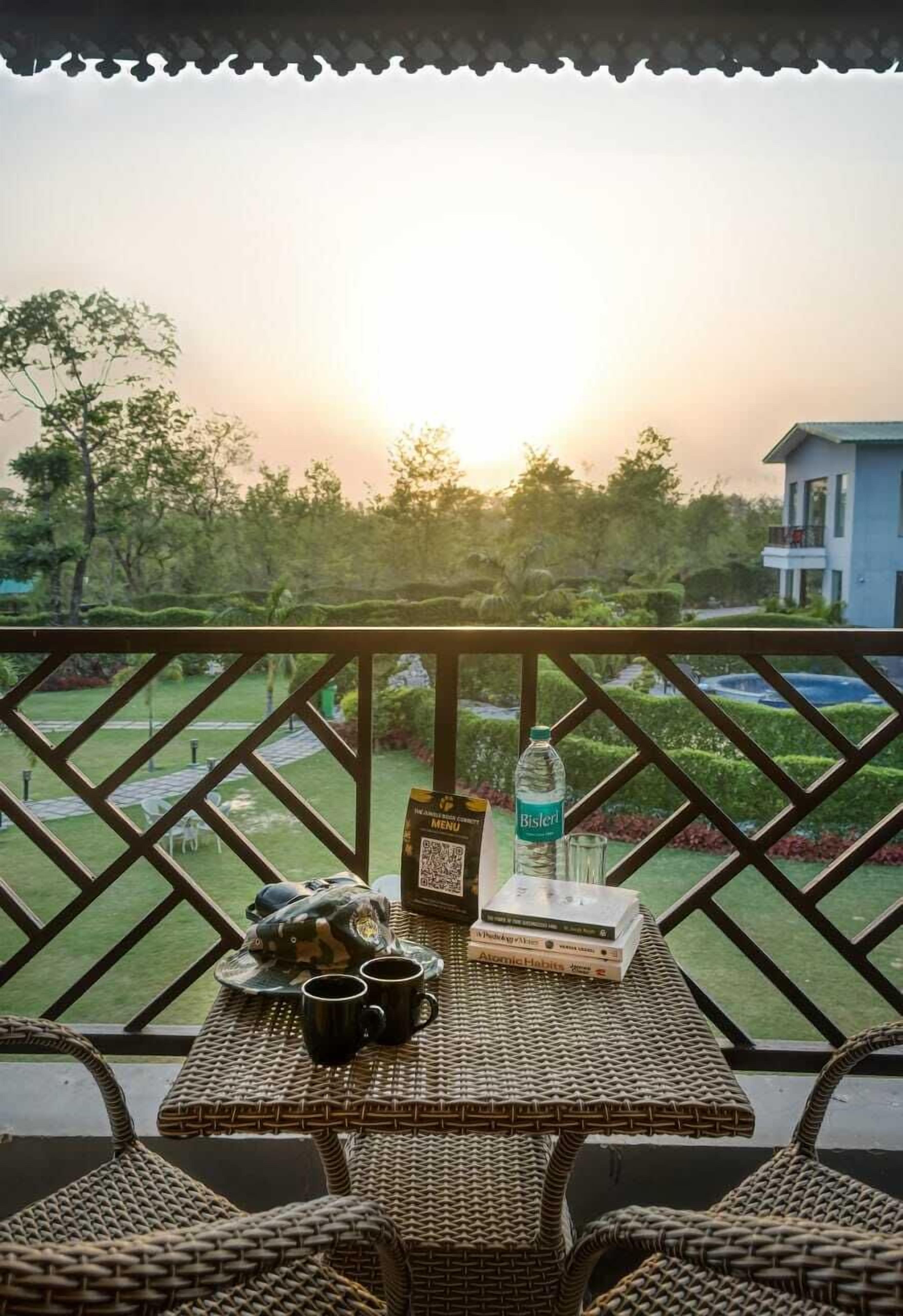 A balcony with wicker furniture, a table with a bottle and books, and a view of a garden and building in the background.