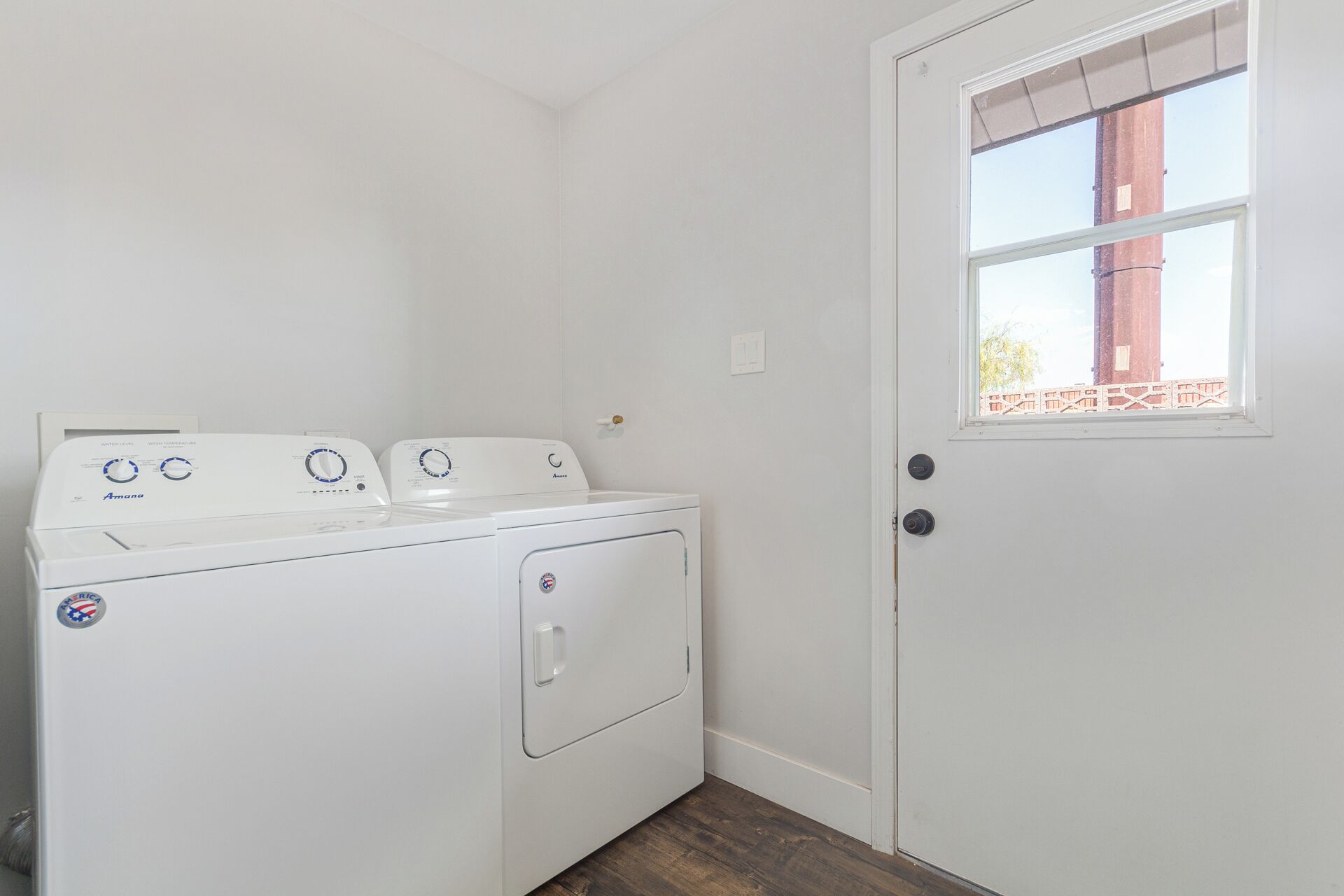 A laundry room with a front-loading washer and dryer, a door with a window, and a wooden floor.