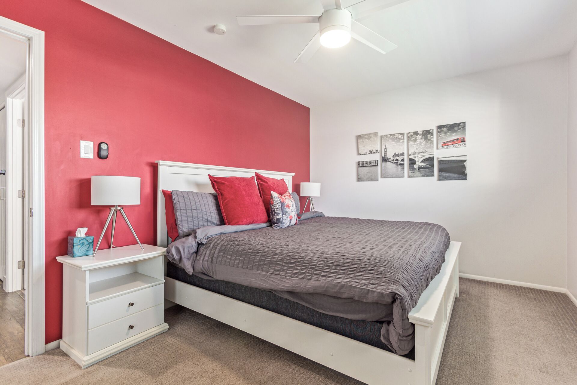 Une chambre avec un lit blanc, des murs rouges et un ventilateur de plafond. Trois tableaux encadrés sont accrochés au mur.