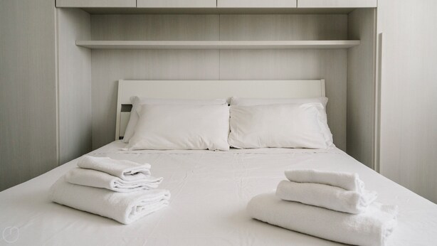 Cama bem arrumada com lençóis e almofadas brancos, e uma toalha dobrada sobre a cama, com uma prateleira embutida acima.
