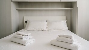 A neatly made bed with white linens and pillows, a folded towel on the bed, and a built-in shelf above the bed. - Mimosa 1 int 7 (Cervo)