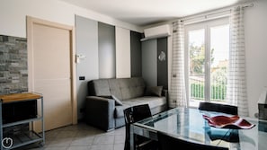 Una sala moderna con un sofá gris, una mesa de comedor de vidrio y una zona de cocina con microondas y un carrito pequeño.