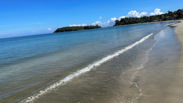 A beach with clear blue water, a sandy shore, and a distant island with vegetation.