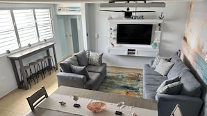 A living room with two grey sofas, a dining table with chairs, a TV mounted on the wall, and a window with blinds.