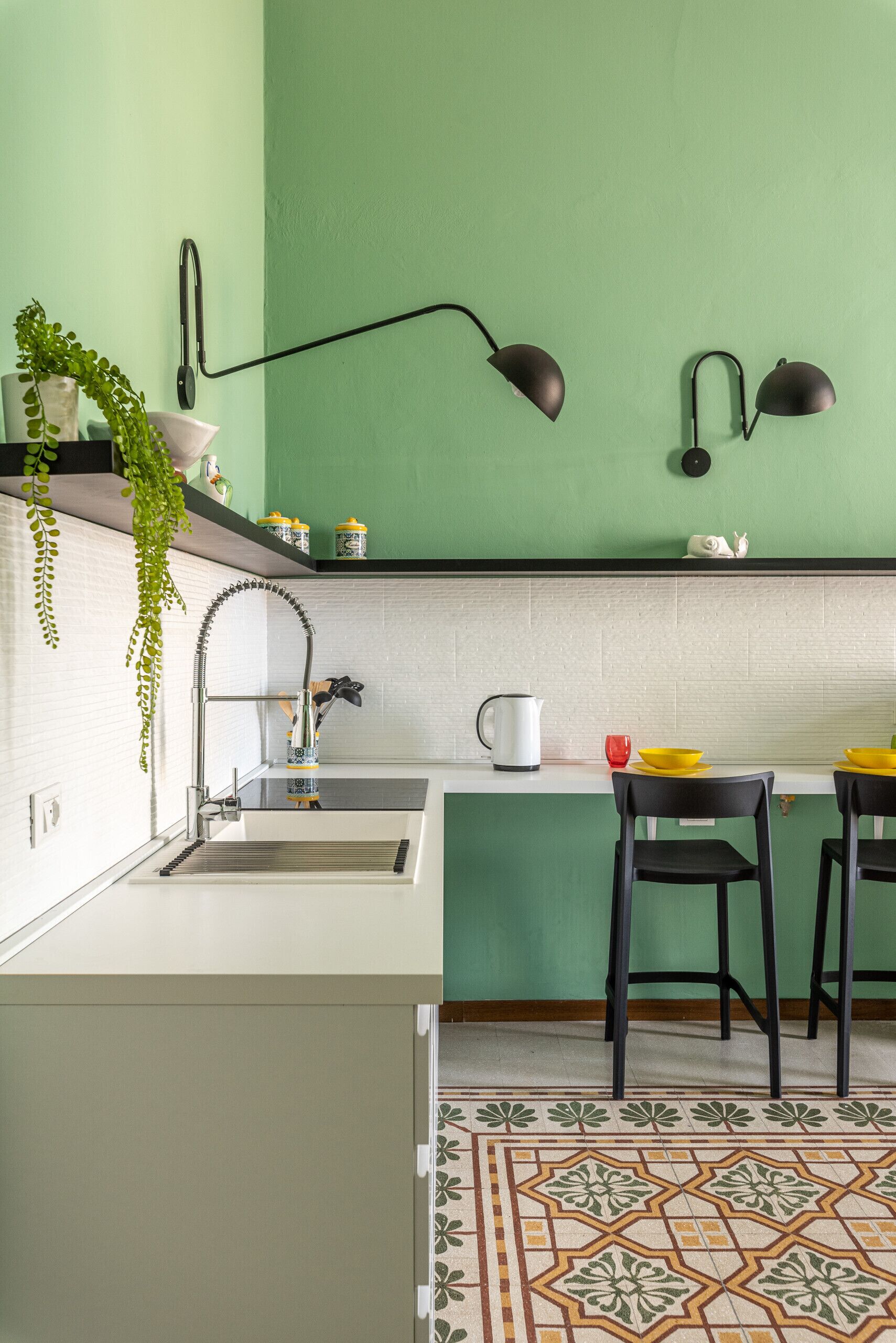 Une cuisine avec un mur vert, des armoires blanches, un évier, deux chaises noires et un sol à motifs.